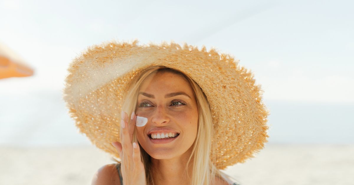 Blonde Frau mit Sonnenhut aus Stroh, cremt sich am Strand das Gesicht ein.