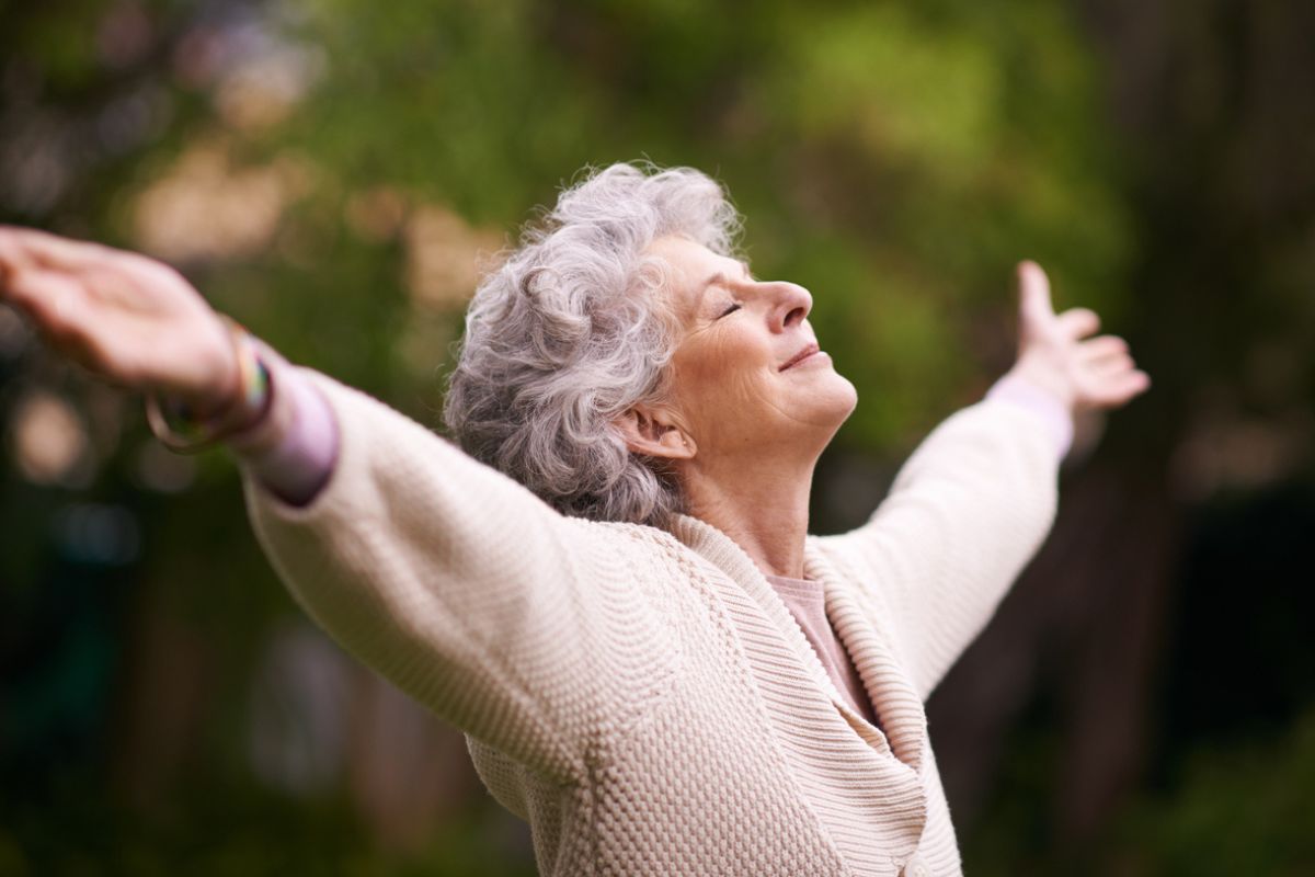 Reife Frau streckt die Arme nach oben, hat die Augen geschlossen und genießt die Natur.